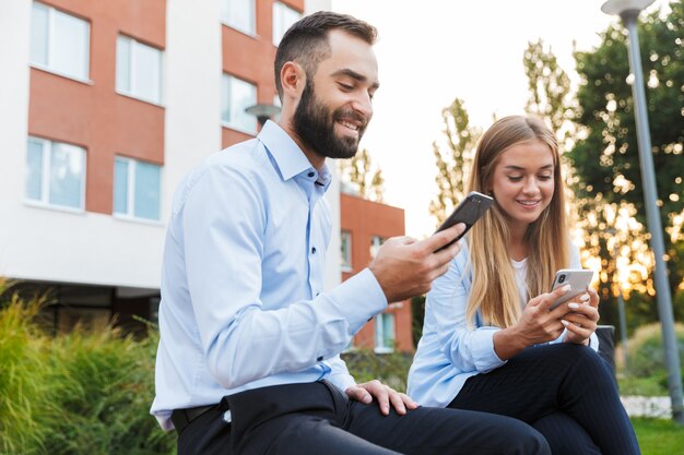 jonge gelukkige collega's man en vrouw zakenmensen zitten buiten op straat met behulp van mobiele telefoons.