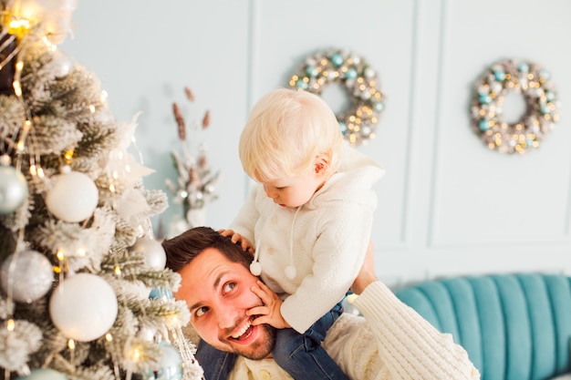 Jonge gelukkige en lachende vader met grappige blonde peuterjongen op zijn schouders in de buurt van versierde kerstboom Oudejaarsavond plezier met familie thuis