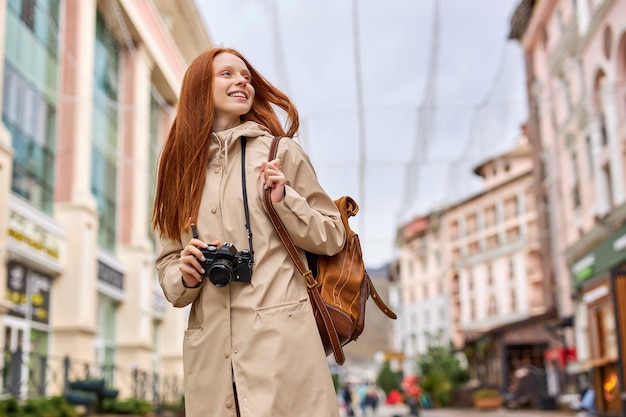 Jonge gelukkige jonge roodharige reiziger vrouw met retro filmcamera in handen, wandelen in een stedelijke straat alleen, beige jas, casual outfit dragen. Leuke blanke dame brengt weekend door in de oude stad?