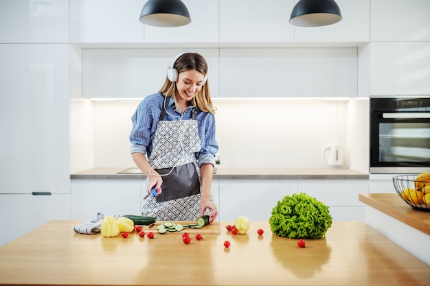 Jonge gelukkige Kaukasische vrouw in schort die zich in keuken bevinden en komkommer snijden terwijl het luisteren van muziek. Op het aanrecht staan allerlei soorten groenten. Gezond eten concept.