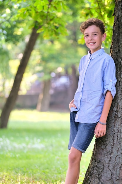 Jonge gelukkige kind jongen ontspannen in zomer park leunend op boomstam Positieve jongere genieten van zomer buiten Kind welzijn concept