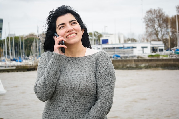 jonge gelukkige latijns-vrouw praten aan de telefoon staande op een pier met de rivier op de achtergrond