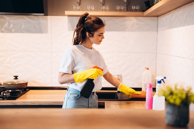 Jonge gelukkige leuke vrouw in gele handschoenen maakt haar keuken thuis schoon met wasmiddelen.