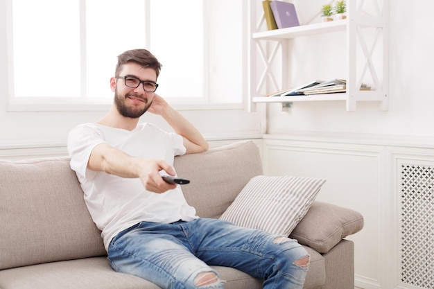 Jonge gelukkige man die tv kijkt op de bank, wijzend met de afstandsbediening op de tv, kopieer ruimte