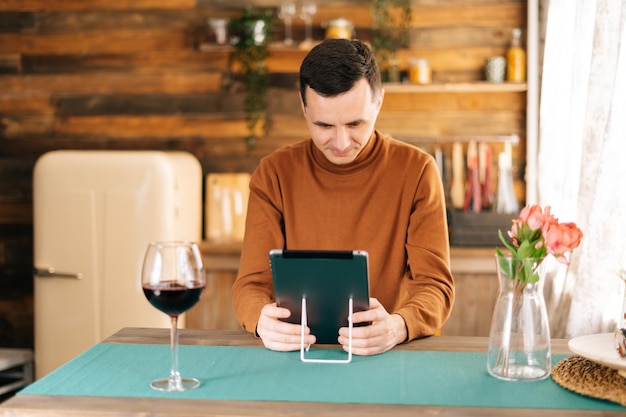 Jonge gelukkige man gebruikt digitale tablet, hoog glas met rode wijn op tafel in een gezellige keukenkamer met houten wanden.