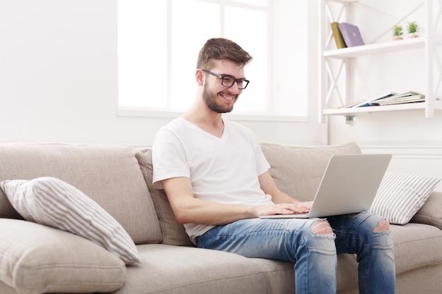 Jonge gelukkige man thuis online chatten op laptop met vriend. Casual man in glas op beige bank in lichte woonkamer, kopieer ruimte