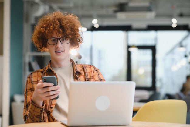 Jonge gelukkige mannelijke freelancer in vrijetijdskleding die in café met laptop zit en mobiele telefoon gebruikt