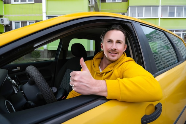 Jonge gelukkige mannelijke taxichauffeur zit achter het stuur van een taxi en laat zien zoals