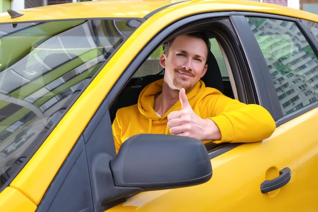 Jonge gelukkige mannelijke taxichauffeur zit achter het stuur van een taxi en shows zoals