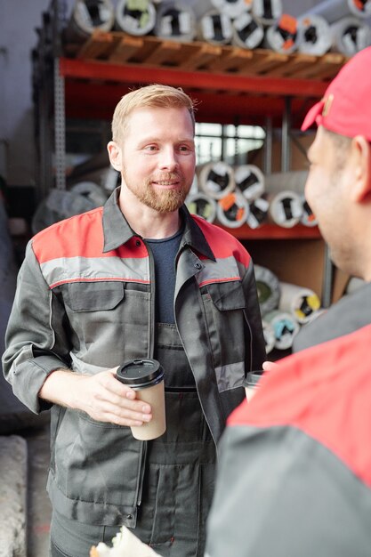 Jonge gelukkige mannelijke werknemer in uniform met koffie op de werkplek tijdens gesprek met collega tijdens lunchpauze tegen rek met goederen