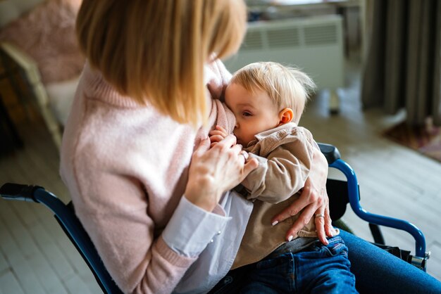 Jonge gelukkige moeder met handicap in rolstoel die haar kind thuis borstvoeding geeft