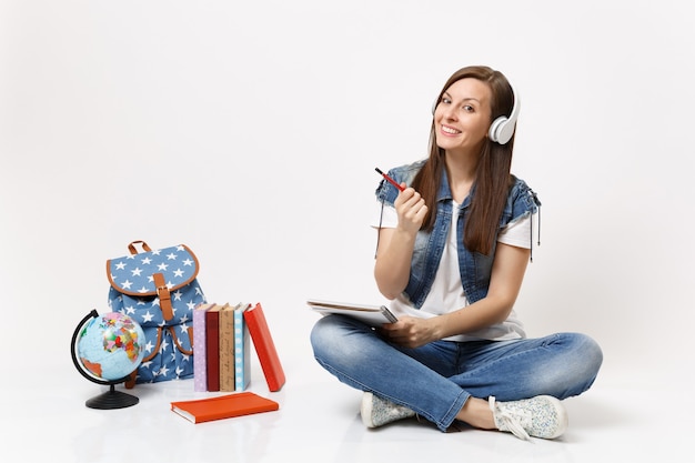Jonge gelukkige mooie vrouw student in koptelefoon luisteren muziek met notebook, potlood in de buurt van globe, rugzak, schoolboeken geïsoleerd op een witte muur