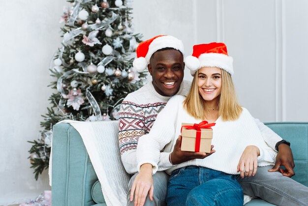 Jonge gelukkige multi-etnisch paar knuffelen en ontspannen op de comfortabele bank. Jonge man en vrouw kijken naar de camera en glimlachen. Portret van een mooi paar in de kersttijd.