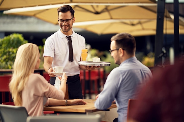 Jonge gelukkige ober die koffie en drankjes brengt voor gasten op een terras