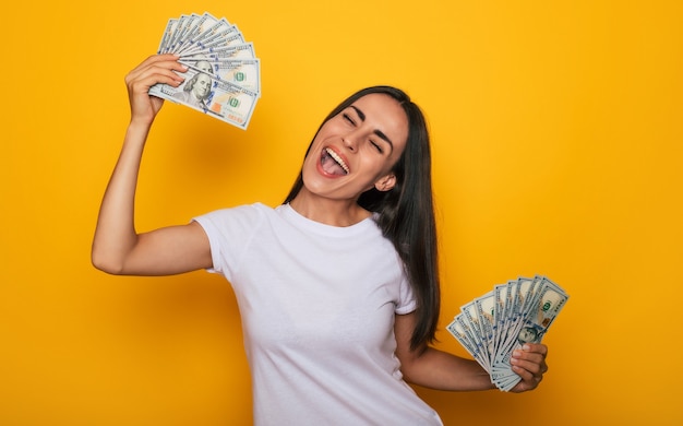 Jonge, gelukkige opgewonden mooie vrouw die emotioneel in de camera kijkt met veel geld in haar handen en plezier heeft geïsoleerd op gele achtergrond