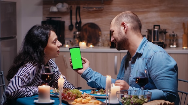 Jonge gelukkige paar met groen scherm telefoon tijdens het diner. Vrolijke mensen kijken naar mockup sjabloon chroma key geïsoleerd smartphone display met behulp van techology internet zittend aan de tafel in de keuken.