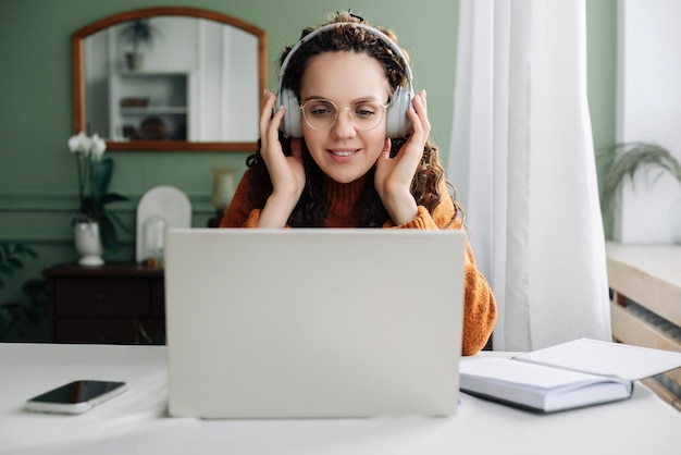 Jonge gelukkige studente die hoofdtelefoon draagt die laptop elearning gebruiken op kantoor aan huis luisterende muziek