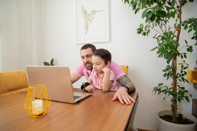 Jonge gelukkige vader in roze poloshirt die op laptop werkt terwijl hij aan tafel zit met zijn zoontje, een gelukkige familie die thuis werkt concept