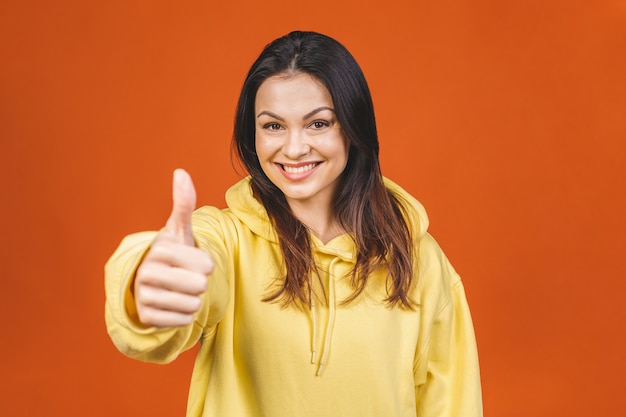 Jonge gelukkige vrolijke vrouw die duimen toont die tegen oranje achtergrond worden geïsoleerd.