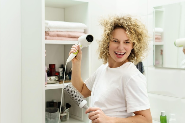 Jonge gelukkige vrouw die een haardroger gebruikt voor haar krullend haar in een badkamer