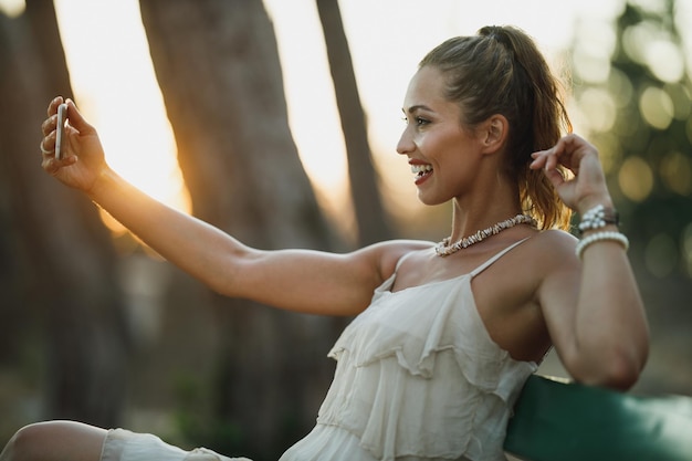 Jonge gelukkige vrouw die een selfie maakt met een smartphone terwijl ze geniet op een bankje in het park tijdens de zomervakantie.