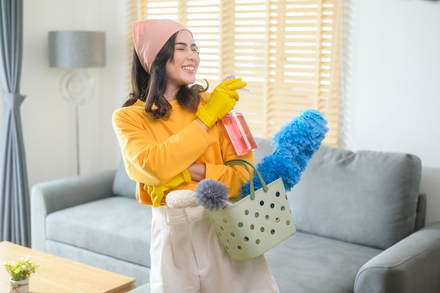 Jonge gelukkige vrouw die gele handschoenen draagt en een mand met schoonmaakspullen in de woonkamer houdt