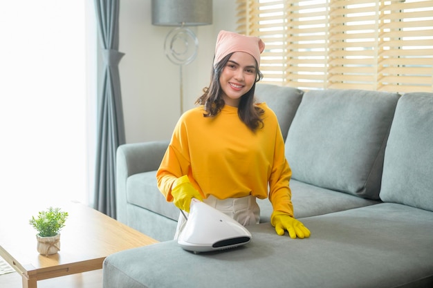 Jonge gelukkige vrouw die gele handschoenen draagt en stofzuigt een bank in de woonkamer schoonmaken