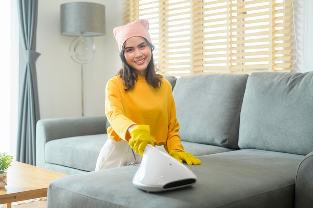 Jonge gelukkige vrouw die gele handschoenen draagt en stofzuigt Een bank in de woonkamer schoonmaken