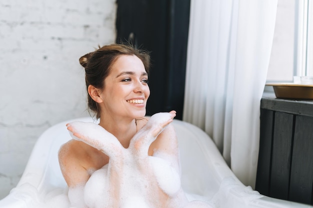 Jonge gelukkige vrouw die thuis een bad met schuim neemt, trakteer uzelf