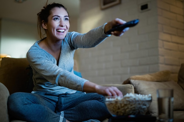 Jonge gelukkige vrouw die van kanaal verandert met afstandsbediening terwijl ze tv kijkt en 's avonds thuis popcorn eet