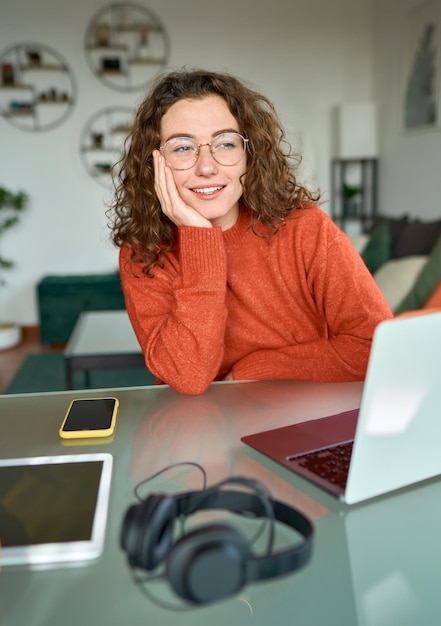 Jonge gelukkige vrouw die wegkijkt en nadenkt terwijl ze thuis aan een laptop werkt