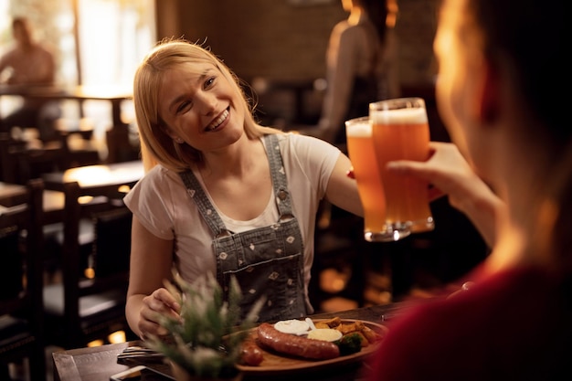 Jonge gelukkige vrouw en haar vriendin eten in een restaurant en hebben plezier tijdens het roosteren met bier