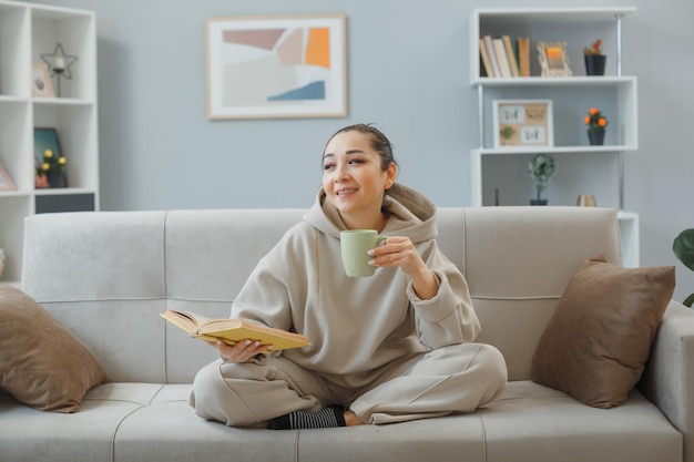 Jonge gelukkige vrouw in casual kleding zittend op een bank in het interieur met een mok die thee drinkt en een boek leest dat er positief uitziet met een glimlach op haar gezicht