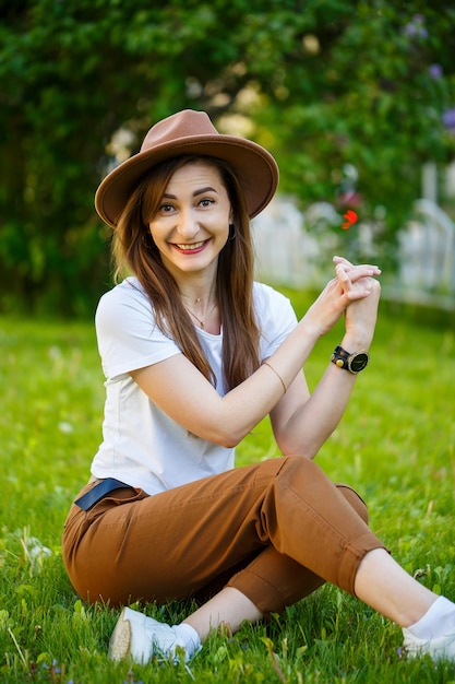 Jonge gelukkige vrouw in een hoed zit op een groen gazon in een park. Een meisje met een Europese uitstraling met een glimlach op haar gezicht op een zonnige zomerdag