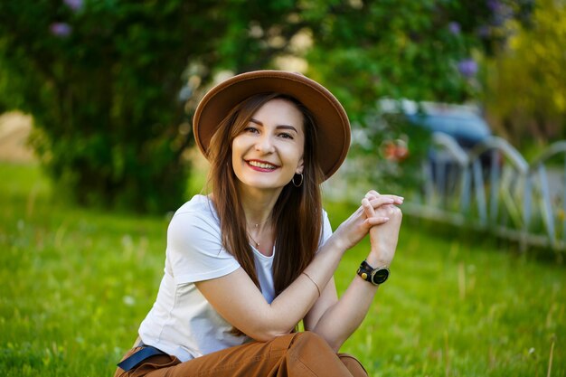 Jonge gelukkige vrouw in een hoed zit op een groen gazon in een park. Een meisje met een Europese uitstraling met een glimlach op haar gezicht op een zonnige zomerdag