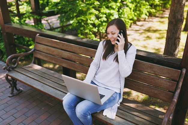 Jonge gelukkige vrouw in lichte casual kleding praten op mobiele telefoon. Vrouw zittend op een bankje bezig met moderne laptop pc-computer in straat buiten op de natuur. Mobiel kantoor. Freelance bedrijfsconcept.