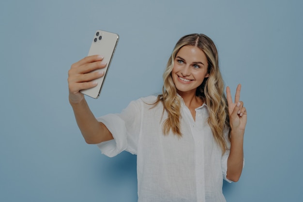 Jonge gelukkige vrouw in vrijetijdskleding die selfie neemt terwijl ze vredesgebaar toont met haar hand, glimlachend met brede glimlach voor volgers en zichzelf uitdrukkend, geïsoleerd over blauwe muur