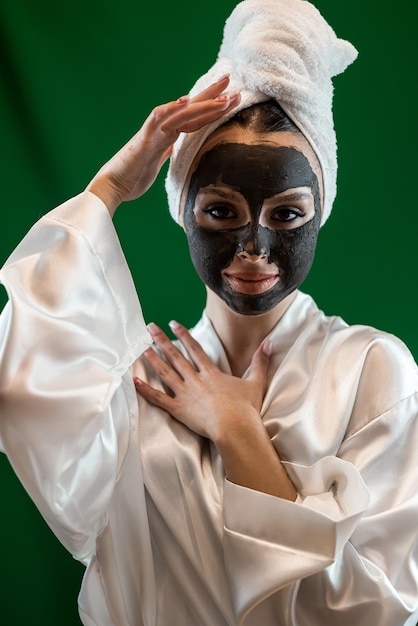 Jonge gelukkige vrouw met een handdoek die therapeutisch voedend zwart kleimasker op de gezichtshuid aanbrengt Procedure voor huidverzorging reinigende spa