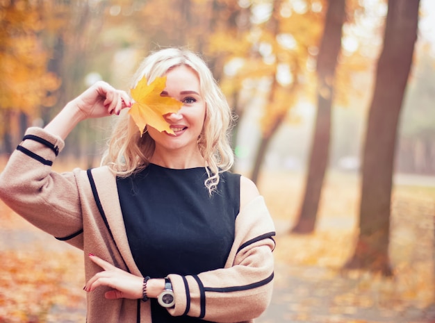 Jonge gelukkige vrouw met geel blad in de buurt van haar gezicht in het herfstpark