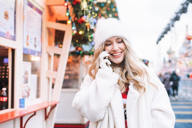 Jonge gelukkige vrouw met krullend haar in witte bontmuts met mobiele telefoon op de kerstmarkt in de winterstraat versierd met lichtjes