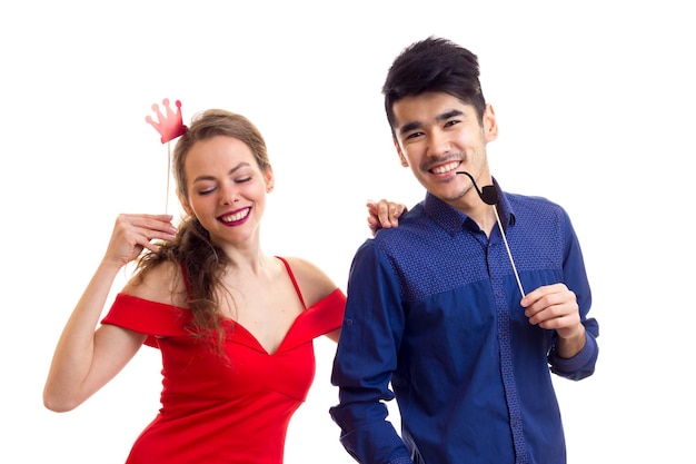 Jonge gelukkige vrouw met lang blond haar in rode jurk en jonge knappe man met donker haar in blauw shirt met kartonnen stokken rookpijp en rode kroon op witte achtergrond in studio.