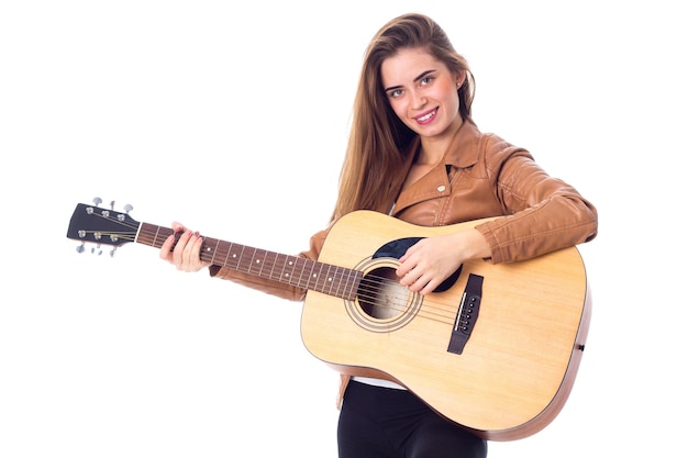 Jonge gelukkige vrouw met lang haar in bruin jasje die een gitaar op witte achtergrond in studio houdt