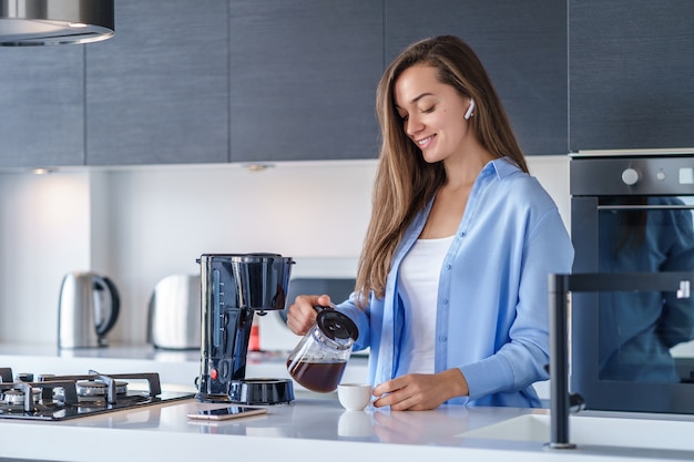 Jonge gelukkige vrouw met witte draadloze oortelefoons het luisteren muziek en audioboek tijdens het maken van verse aromatische koffie thuis gebruikend koffiezetapparaat in de keuken. Moderne mobiele mensen