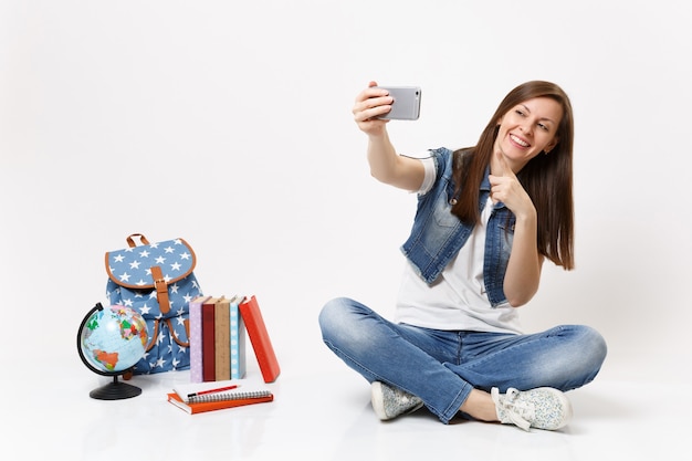 Jonge gelukkige vrouw student doen selfie schot op mobiele telefoon, wijzende wijsvinger in de buurt van globe, rugzak, schoolboeken geïsoleerd