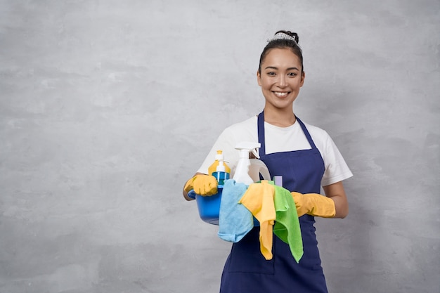 Jonge gelukkige vrouw vrouwelijke reiniger in uniform en rubberen handschoenen met emmer schoonmaakbenodigdheden