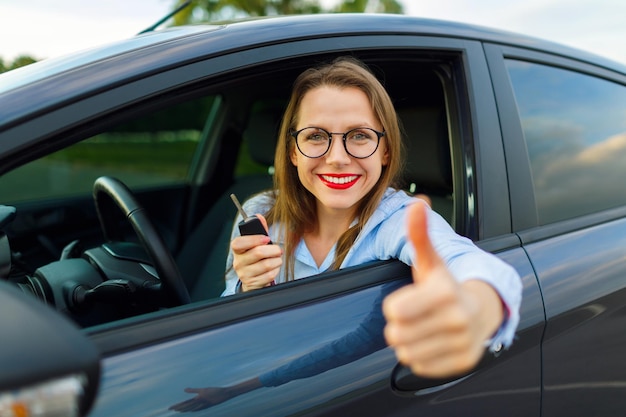 Foto jonge gelukkige vrouw zit in een auto met de duim omhoog concept van het kopen van een gebruikte auto of een gehuurde auto