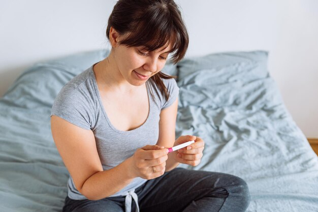 Jonge gelukkige vrouw zit op bed en kijkt naar een positieve zwangerschapstest.