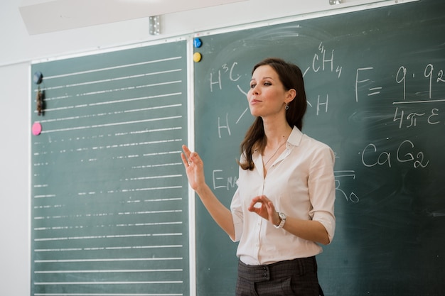 Jonge gelukkige vrouwelijke leraar die aan klasse spreekt die zich tegen groene raad bevindt