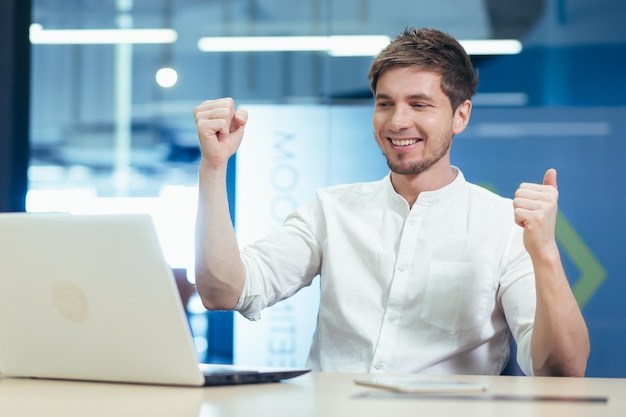 Jonge gelukkige zakenman kijkt naar laptopscherm houdt handen omhoog met vreugde viert triomf overwinningsgebaar man aan het werk op kantoor