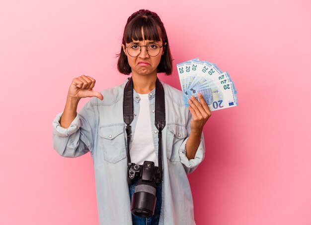 Jonge gemengd ras fotograaf vrouw met rekeningen geïsoleerd op roze achtergrond met een afkeer gebaar, duim omlaag. Onenigheid begrip.